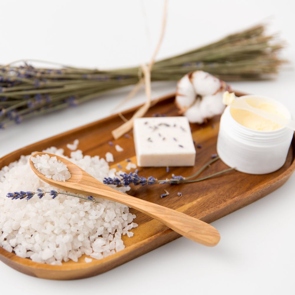 Wooden tray with sea salt, lavender flowers, a wooden spoon, soap, and a small container on a white background.