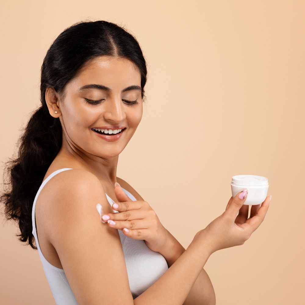 Woman applying cream to her shoulder with a beige background
