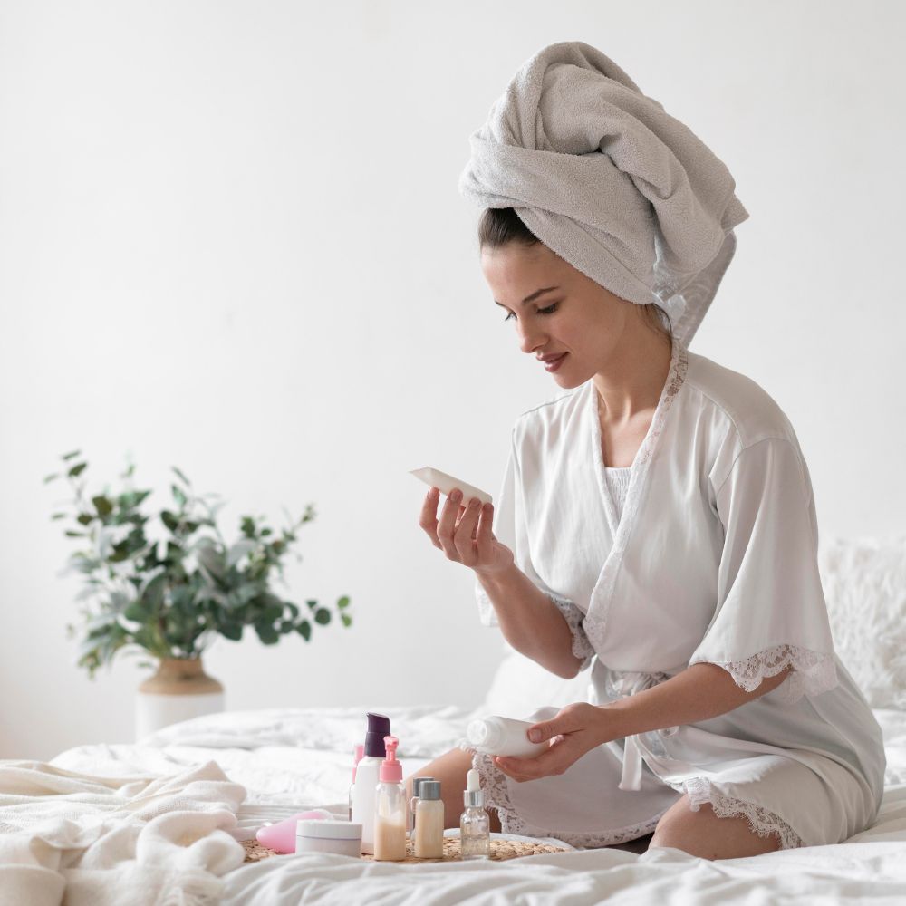 Woman in a white robe with a towel on her head, sitting on a bed with skincare products around her.